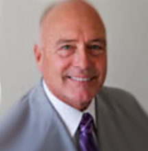 Image of Robert Zumberge; white male with blue eyes, bald head with some white hair in the back, blue eyes, wearing a gray suit with purple tie.